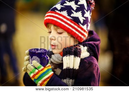 Little cute kid boy with cup of steaming hot chocolate or children punch. Happy child on Christmas market in Germany. Preschool kid in winter clothes warming hands on cup and drinking eggnog
