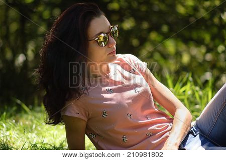 Outdoors Portraiit of Young Woman in Fashionable Sunglasses and Pink Shirt Lying on the Ground During Sunny Day in the Summer