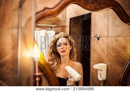 beautiful girl blows with a hair dryer and looks at her reflection in the bathroom mirror. She is going on a date
