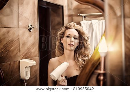 beautiful girl blows with a hair dryer and looks at her reflection in the bathroom mirror. She is going on a date