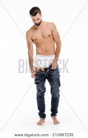 Getting undressed. Full length of handsome shirtless young man taking off his jeans and looking away while standing against white background.