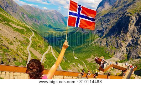 Tourist Woman With Norwegian Flag On Trollstigen Viewpoint. People On Viewing Platform In The Backgr