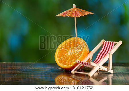 A Small Chaise Longue And Half An Orange With A Cocktail Umbrella On Wet Boards, Against The Backgro