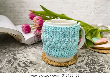 White Cup Of Tea In A Knitted Blue Case With An Open Book And With Pink Flowers Tulips And Sweets, W