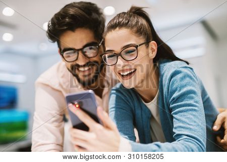 Two Beautiful Happy Young People Checking Out The Mobile Specifications While Shopping In The Electr