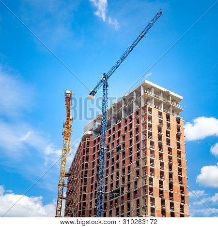 Construction Site With Cranes Near The Building Against The Blue Sky With White Clouds. Construction