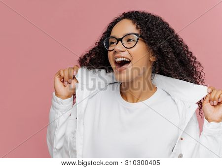 Curly Overemotive Woman Laughs Happily, Keeps Hands On Collar Of White Jacket, Has Healthy Skin, Wea