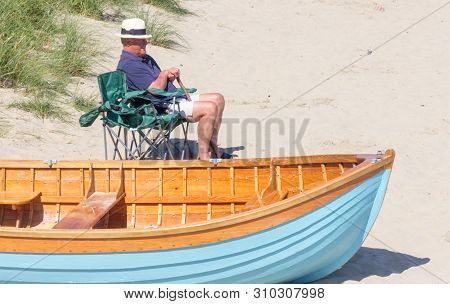 Porth Nefyn, Wales - August 18th 2015: Senior Man Sat In Chair On The Beach Next To A Boat. People F