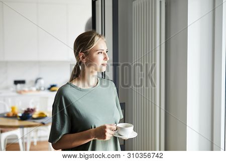 Young Casual Woman With Cup Of Coffee Looking Through Window While Staying At Home On Weekend