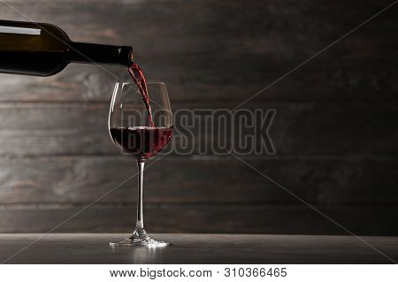 Pouring Red Wine Into Glass From Bottle On Table Against Wooden Background. Space For Text