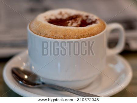 Selective Focus On Cappuccino On Table At A Cafe Filling The Frame, With Newspapers In The Backgroun
