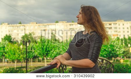 Content Woman Looking Dreamily Standing In Green Park Against Ornamental Iron Fence. Shy Woman Leani