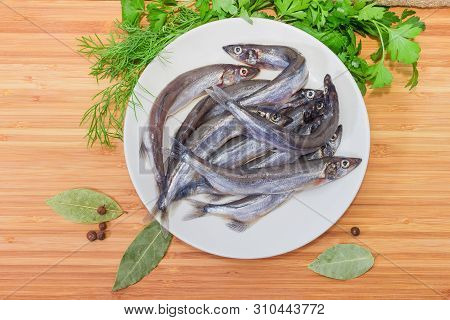Whole Defrosted Uncooked Capelin On Dish Among Some Greens And Spices On The Wooden Surface, Top Vie
