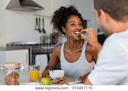 Man feeding cookie biscuit to his girlfriend during breakfast. Happy boyfriend feeding his girl with homemade cookie in kitchen. Closeup of multiethnic couple eating breakfast at home. 