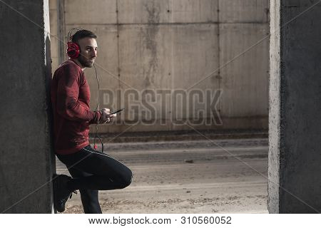 Muscular Athetic Man Wearing Sportswear, Leaning Onto A Concrete Wall, Choosing A Song On A Smart Ph