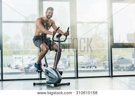 Smiling Handsome Sportsman With Electrodes On Elliptical During Endurance Test In Gym