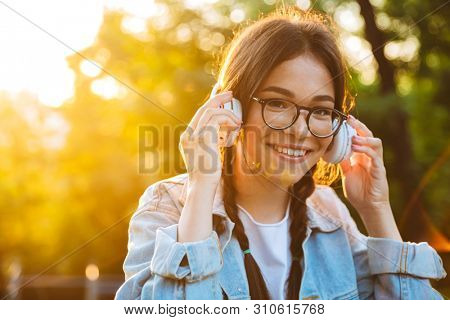 Image of a optimistic smiling young teenage girl student sitting outdoors in beautiful green park listening music with headphones.