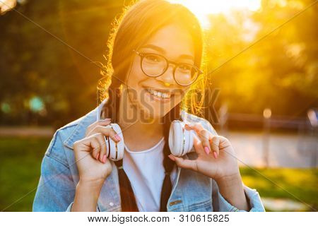Image of a pleased happy young teenage girl student sitting outdoors in beautiful green park listening music with headphones.