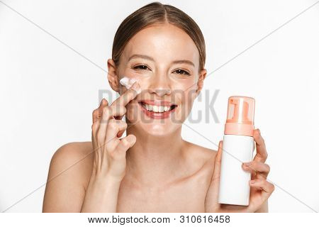 Image of beautiful half-naked woman smiling while washing her face with foaming facial cleanser isolated over beige background