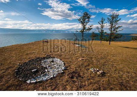 Yin-yang Sign Of Colored Stones On The Shore Of Lake Baikal. Trees On The Steppe Shore. Behind The M