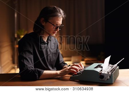 Profile Of A Sad Man In Eyeglass, Seated At A Wooden Table, Working On A Old Vintage Typewriter In D