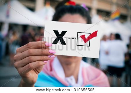 closeup of a young person, wrapped in a transgender pride flag, holding a piece of paper with a check mark on an X letter for the third gender category