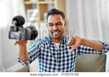 blogging, videoblog and people concept - displeased indian male video blogger with camera videoblogging and showing thumbs down at home