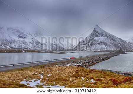Lofoten Islands In Norway. Red Noticable Car Passing By Fredvang Bridge At Lofoten Islands In Winter
