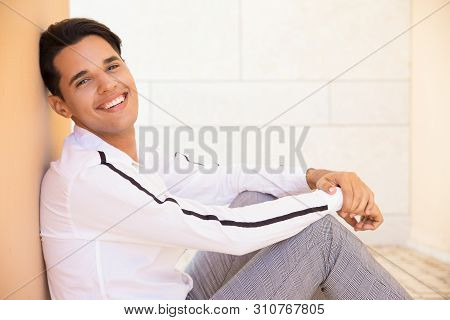 Happy Latin Man Sitting And Reclining On Wall Outdoors. Handsome Young Man Wearing Shirt, Looking At