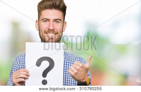 Young handsome man holding paper with question mark happy with big smile doing ok sign, thumb up with fingers, excellent sign