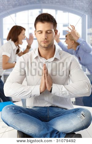 Forretningsmand meditere på kontor med øjnene lukkede, sidder på skrivebord, kolleger argumenterer i baggrunden.