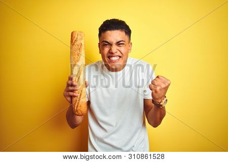 Young brazilian baker man holding bread standing over isolated yellow background screaming proud and celebrating victory and success very excited, cheering emotion
