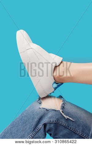Trendy White Eco Leather Sheakers On Bent Leg Knee In Ragged Jeans Over Pastel Turquoise Blue Backgr