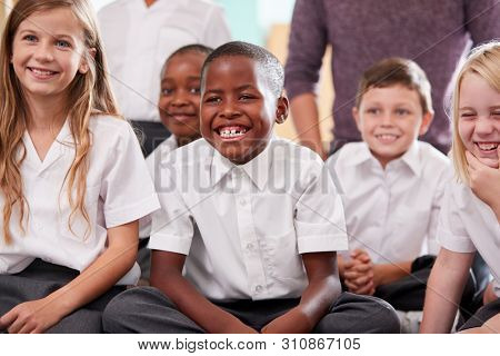 Group Of Elementary School Pupils Wearing Uniform Sitting On Floor Listening To Teacher
