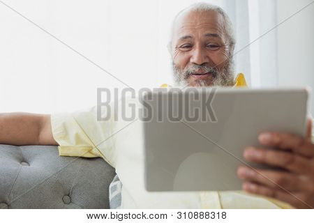 Front view of mixed race man using digital tablet sitting on a couch. Authentic Senior Retired Life Concept