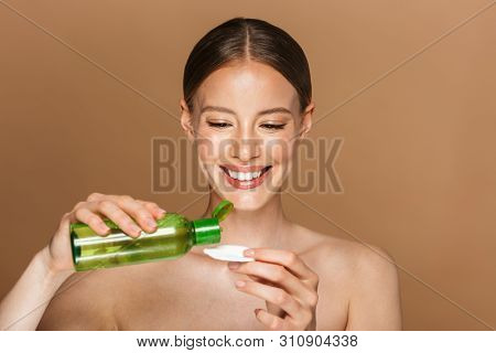 Beauty portrait of an attractive smiling blonde topless woman standing isolated over beige background, cleancing her face with tonic and cotton pad