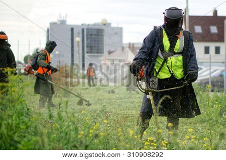 Men In Protective Suits Mow The Grass On A City Lawn With Manual Diesel Lawn Mowers. Improvement And