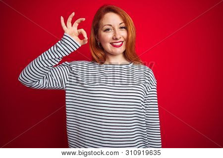 Young redhead woman wearing strapes navy shirt standing over red isolated background smiling positive doing ok sign with hand and fingers. Successful expression.