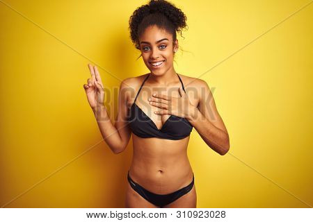 African american woman on vacation wearing bikini standing over isolated yellow background smiling swearing with hand on chest and fingers up, making a loyalty promise oath