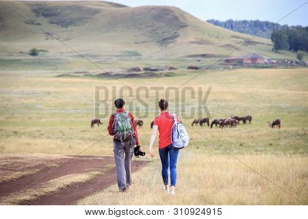 Two Young Girls With Backpacks And Photo Camera On Rural Road. Horse Farm Pasture With Mare And Foal