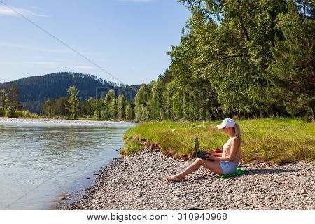 A Young Beautiful Woman Blonde In A White Cap And Denim Shorts Sits On The Rocky Shore Of The River 