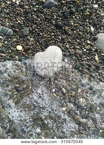 large rounded light grey stone in shape of heart lies under the water near the shore, a running wave, glare on the water, beach pebbles of small colored stones, festive dreamy mood, relax atmosphere, there is free space for writing, copispace, Valentine's
