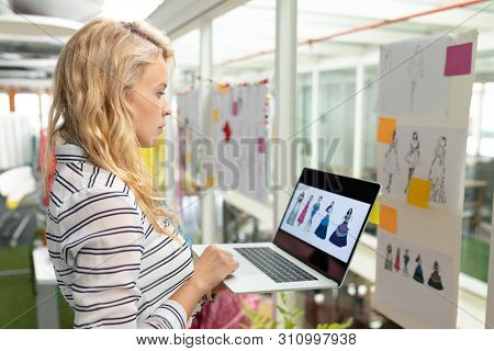 Side view of young Caucasian female graphic designer using laptop in design studio. This is a casual creative start-up business office for a diverse team