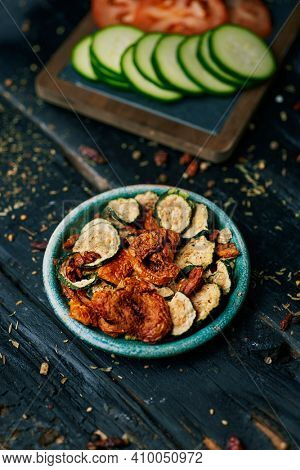 some slices of dried tomato and zucchini, served as appetizer or snack in a rustic green plate, and some slices of raw tomato and zucchini, on a slate tray, placed on a dark gray rustic wooden surface