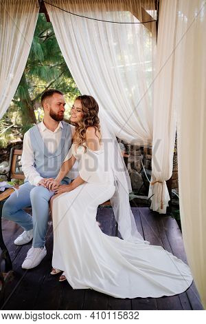 Groom In Blue Suit And Bride With Long Curls With Bouquet Sit In A Gazebo