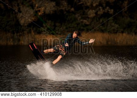Sporty Man In Wetsuit Holds Taut Rope And Rides Wakeboard On The Water Surface