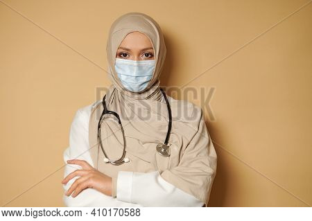 Muslim Female Doctor In Hijab And Medical Blue Mask Looking At Camera With Crossed Arms. Beige Backg