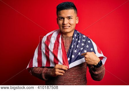 Young handsome latin patriotic man wearing united states of america falg with surprise face pointing finger to himself