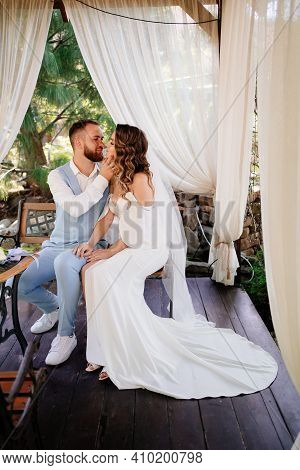 Groom In Blue Suit And Bride With Long Curls With Bouquet Sit In A Gazebo