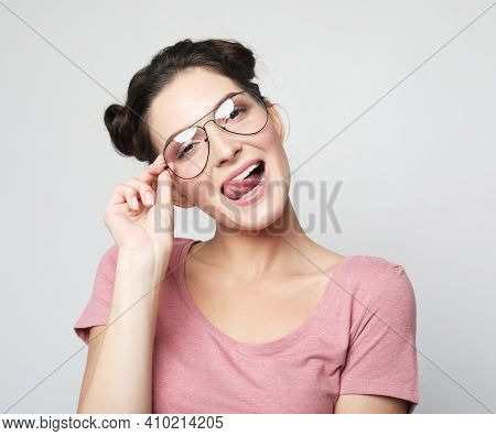 Funny brunette woman wearing pink thirt and glasses showing tongue and looking at the camera over grey background